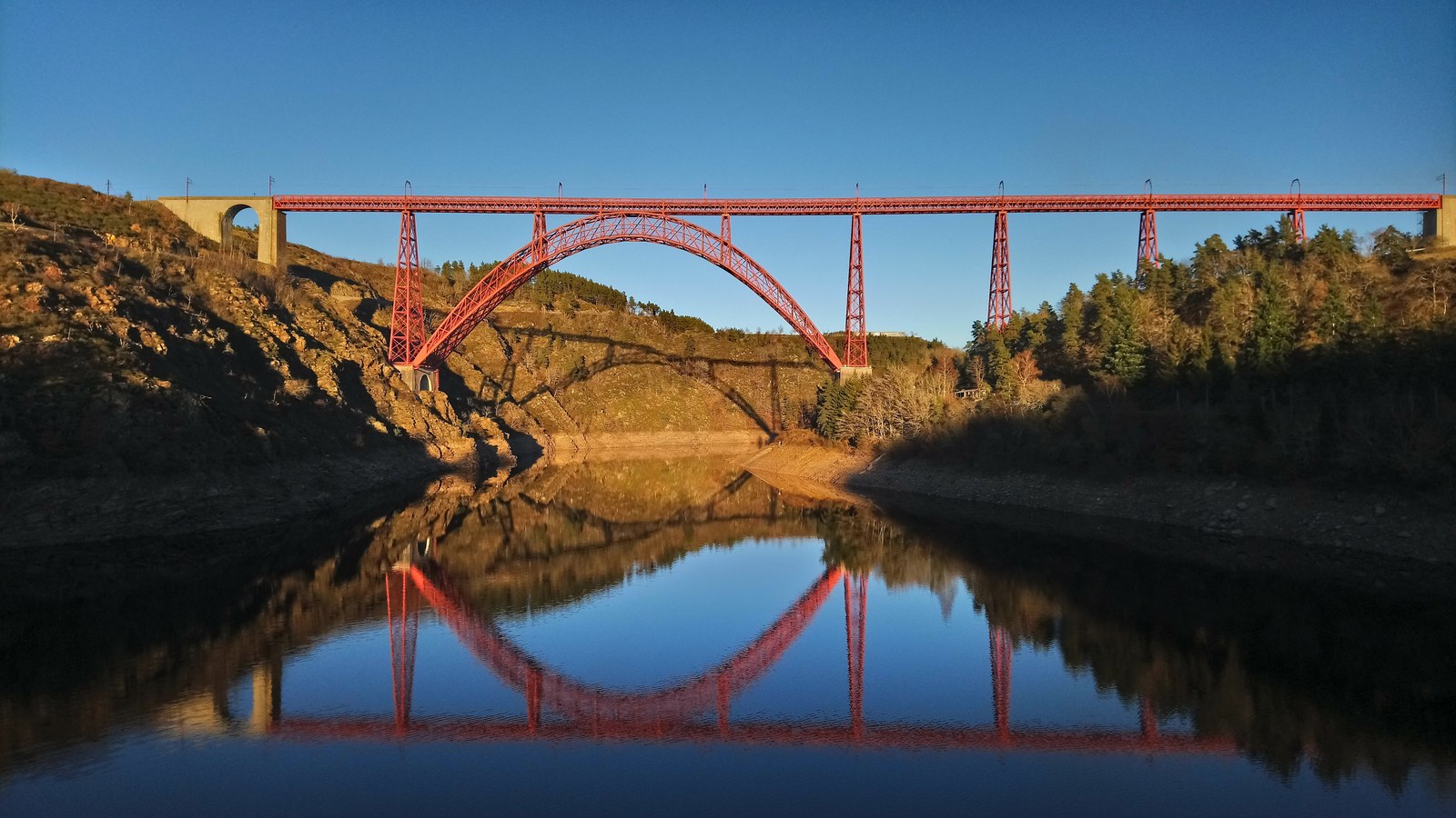 Le viaduc de Garabit, pont ferroviaire en fer rouge à grand arc, se reflète dans le lac de retenue de Grandval au soleil couchant
