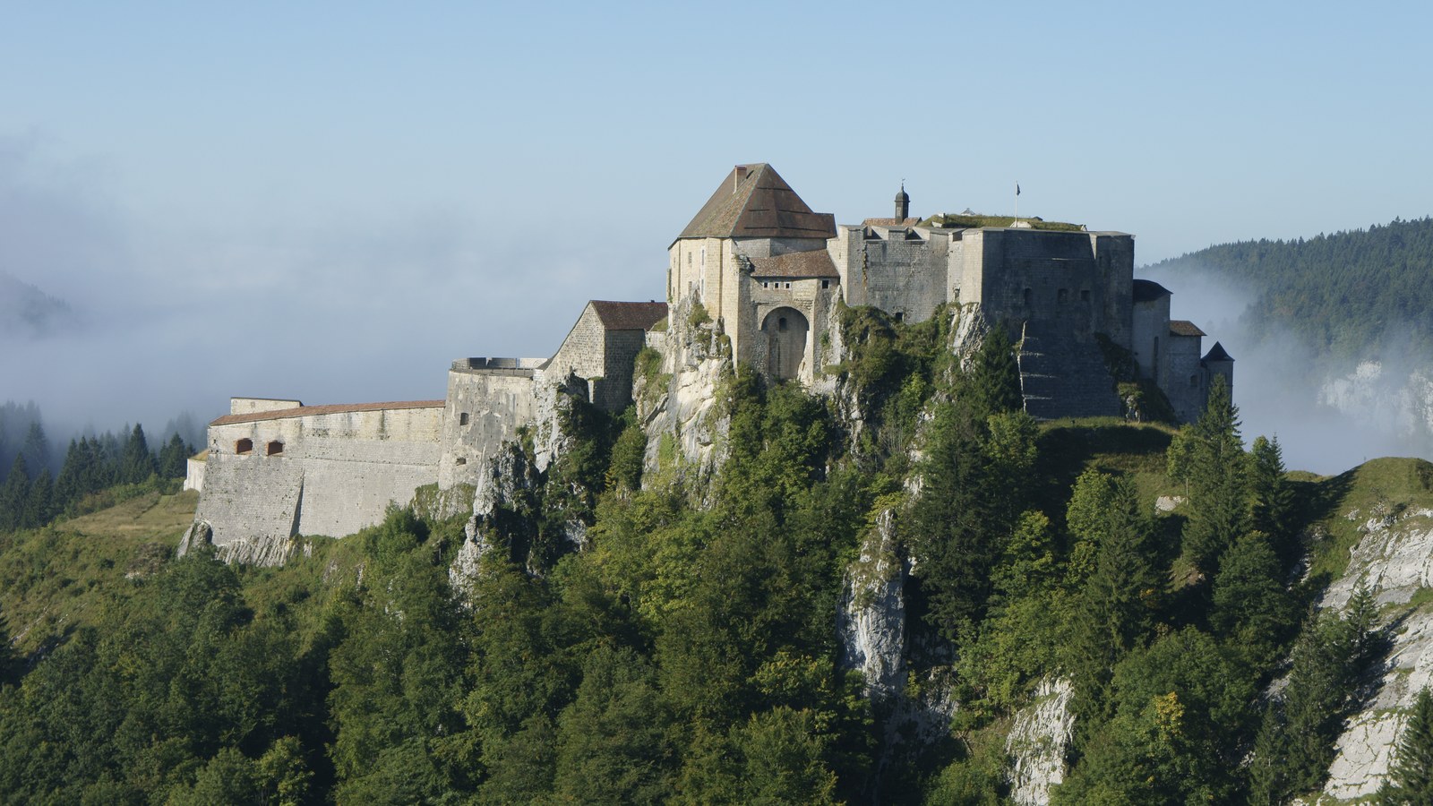 Le château de Joux perché sur son éperon rocheux boisé, avec son donjon et ses enceintes de pierre, au-dessus d'une nappe de brume matinale