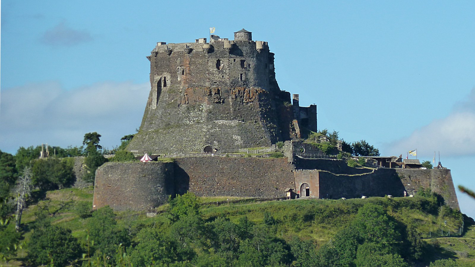 Le château de Murol sur sa butte boisée, avec son enceinte basse au premier plan et la haute forteresse sombre au sommet, sous un ciel bleu