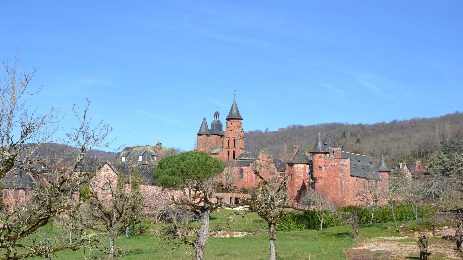 Maisons et tours en grès rouge de Collonges-la-Rouge sous un ciel bleu, avec un pré et des arbres fruitiers au premier plan