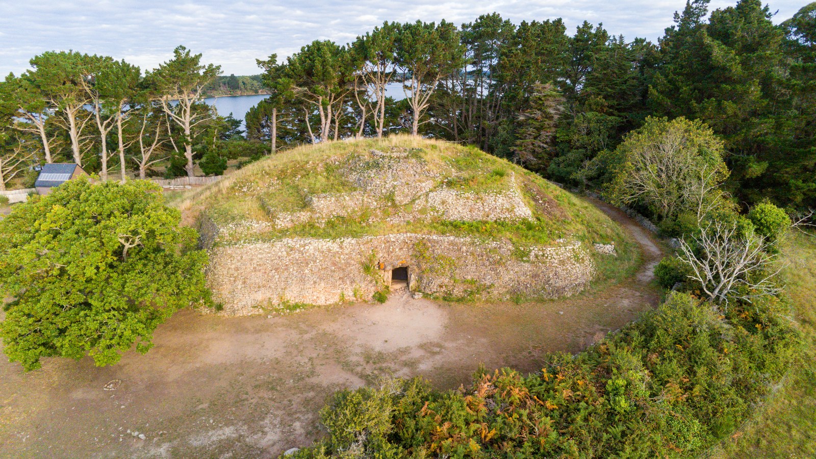 Vue aérienne du cairn de Gavrinis, sa façade en pierres sèches et l'entrée du couloir, entourés de pins au bord du golfe du Morbihan