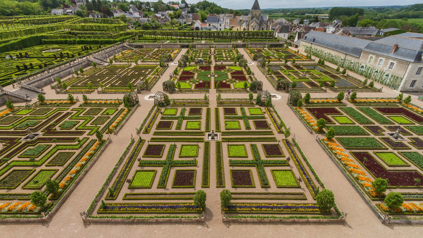 Vue plongeante sur le potager décoratif de Villandry : carrés de salades vertes et rouges bordés de buis, allées de gravier, tonnelles et église du village au fond